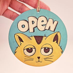 Peut inclure: Panneau rond en bois avec un fond bleu clair et le mot "OPEN" en lettres blanches. En dessous, un visage de chat jaune avec des détails bruns et une expression endormie. Une ficelle marron est attachée pour l'accrocher.