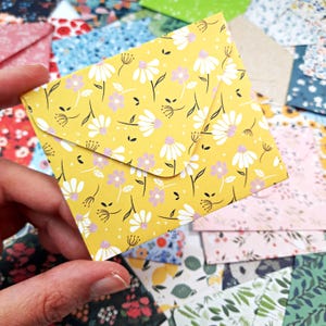 Op de afbeelding: Een kleine, gele bloemenenvelop met een flap, vastgehouden in een hand. De envelop heeft een herhalend patroon van witte madeliefjes, paarse bloemen en zwarte stelen. Andere enveloppen met patronen zijn op de achtergrond.