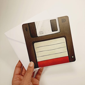 Könnte beinhalten: Eine Grußkarte mit einem Retro-Disketten-Design. Die Karte hat einen braunen Hintergrund, einen roten Streifen und einen weißen Etikettenbereich. Die Karte wird in einer Hand gehalten, mit einem weißen Umschlag sichtbar.