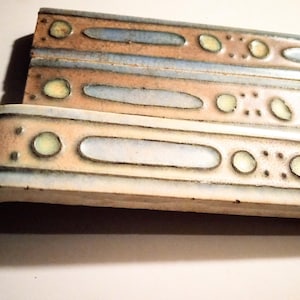 Puede incluir: Tres azulejos de cerámica con un patrón decorativo de azul claro, beige y blanco. Los azulejos tienen un borde redondeado y una superficie texturizada.