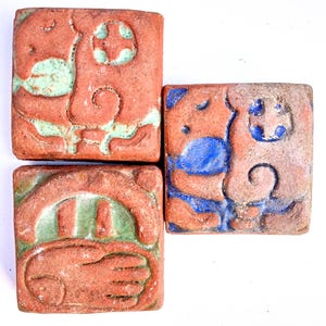 May include: Four square ceramic tiles with different designs. Each tile has a different color scheme, including green, blue, and brown. The tiles are decorated with abstract patterns and designs.