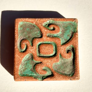 May include: A square terracotta tile with a raised design of abstract shapes in a green glaze.