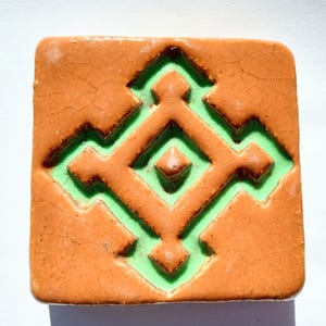 May include: A terracotta tile with a green geometric design. The design is raised and has a diamond shape in the centre.