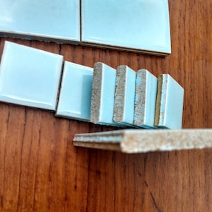 Könnte beinhalten: Ein Stapel hellblauer Keramikfliesen mit einer rauen, unfertigen Kante. Die Fliesen sind in einer Reihe auf einer Holzoberfläche angeordnet.