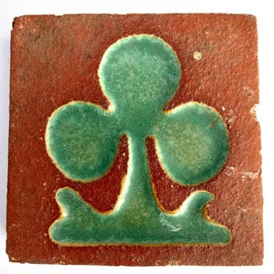 May include: Square, antique tile with a green clover design on a reddish-brown background. The clover is raised and has a glossy finish. The tile has a worn, aged appearance, suggesting it is vintage or reclaimed.