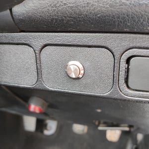 May include: Close-up of a car dashboard with a silver button. The button is centered on a textured, dark gray panel. Other sections of the dashboard are visible, including a red and white button and other compartments.
