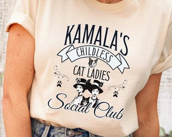 May include: A beige t-shirt with a black graphic design that says "Kamala's Childless Cat Ladies Social Club". The graphic features two women wearing hats and holding cats.