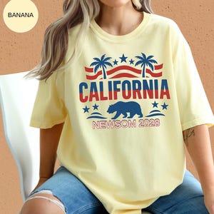 May include: A light yellow t-shirt with a graphic that says "CALIFORNIA" in red and blue, with palm trees, stars, and a bear. The text "NEWSOM 2028" is below the bear. The shirt is a casual style.