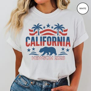 May include: White t-shirt with a graphic design featuring the word "CALIFORNIA" in red and blue, palm trees, stars, and a blue bear. The text "NEWSOM 2028" is below the bear.