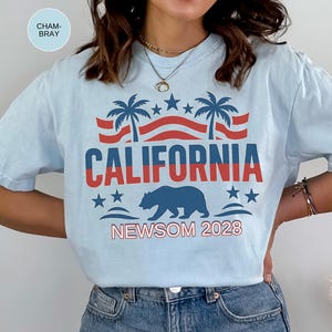 May include: Light blue t-shirt with a graphic design featuring the word "CALIFORNIA" in red and blue, palm trees, stars, and a bear silhouette. The text "NEWSOM 2028" is also included.