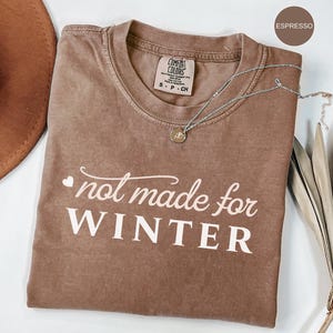 May include: Espresso-colored t-shirt with the text "not made for WINTER" in white script. The shirt has a crew neck and short sleeves. A silver necklace with a small pendant is draped across the shirt. A brown hat is partially visible.