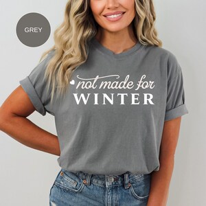 May include: Grey t-shirt with rolled sleeves and the text "not made for WINTER" in white script. The shirt is paired with blue denim jeans. A grey circle with the word "GREY" is in the upper left corner.