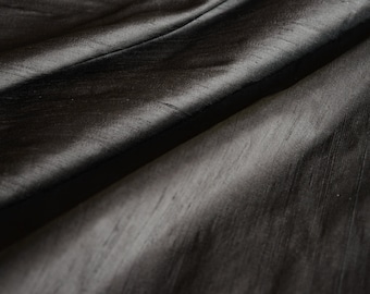 May include: Close-up of a dark gray fabric with a subtle sheen. The material is draped, creating soft folds and shadows. The texture appears smooth with visible, fine lines. The overall effect is elegant and luxurious, suitable for apparel or home decor.