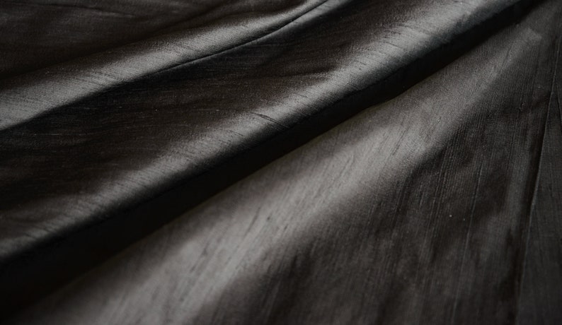 May include: Close-up of a dark gray fabric with a subtle sheen. The material is draped, creating soft folds and shadows. The texture appears smooth with visible, fine lines. The overall effect is elegant and luxurious, suitable for apparel or home decor.