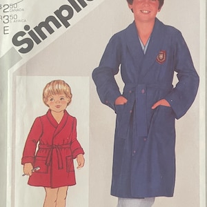 May include: Simplicity pattern envelope showing illustrations of children's robes. The main image depicts a boy in a blue robe with a belt and pockets. A smaller image shows a child in a red robe. The envelope includes the brand name "Simplicity" and sizing details.