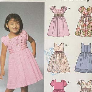 Puede incluir: Un sobre de patrón de costura Simplicity 5704 para talla A 3,4,5,6,7,8 con 6 vestidos fáciles de hacer. El frente muestra a un niño con un vestido de cuadros vichy rosa con mangas abullonadas y bordados florales, junto a seis diseños de vestidos.