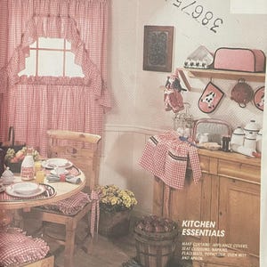 Può includere: Una busta di modello McCall's Crafts con una scena di cucina. Tendine, cuscini e grembiule a quadri rossi e bianchi. Il testo include "Kitchen Essentials" e "Les indispensables de la cuisine".