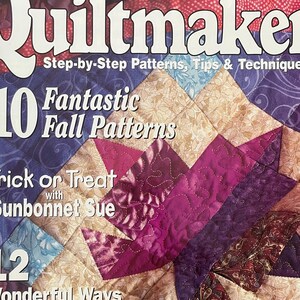 Op de afbeelding: Een tijdschriftcover voor Quiltmaker, een publicatie voor quilters. De cover toont een kleurrijk quilt met een patchwork-ontwerp in tinten paars, roze, blauw en beige. De tekst op de cover luidt "10 Fantastic Fall Patterns" en "Trick or Treat with Sunbonnet Sue".