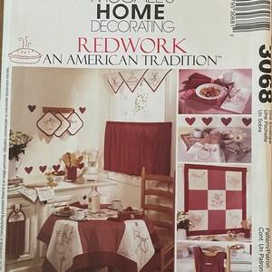 Könnte beinhalten: Ein McCall's Home Decorating-Muster für Rotwerkm Stickerei. Das Muster enthält Anleitungen zum Nähen eines Tischläufers, Platzsets, einer Gardine, einer Teekanne und einer Schürze. Das Muster zeigt ein rotes und weißes Farbschema mit einem Motiv aus Äpfeln und Blättern.