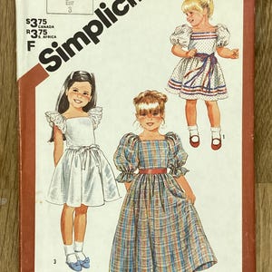 Pode incluir: Um envelope de padrão de costura Simplicity vintage, com ilustrações de três meninas em vestidos. O envelope está rotulado "6062" e inclui texto em inglês e francês. Os vestidos são de vários estilos, incluindo um vestido xadrez.