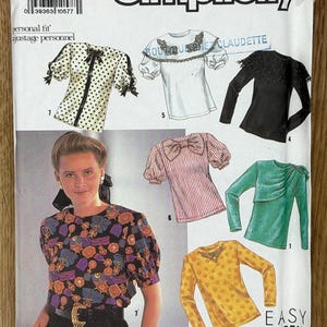 May include: A vintage Simplicity sewing pattern envelope showcasing different blouse designs. The envelope shows illustrations of blouses with details such as polka dots, bows, and lace. The pattern size is N5 (10-18).