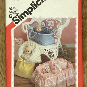Könnte beinhalten: Simplicity-Schnittmuster 6056 für Puppenzubehör. Das Muster enthält Anleitungen für eine Puppentrage, eine Wiege und einen Schlafsack. Das Bild zeigt die fertigen Produkte, darunter eine blaue Denim-Trage, eine rosa Wiege und ein gelber Schlafsack.