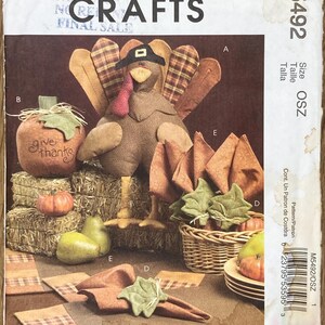 May include: McCall's Crafts pattern M5492 featuring Thanksgiving-themed decorations. The image showcases a stuffed turkey, pumpkin, pears, and napkins. The text "Give thanks" is visible on a pumpkin. The pattern includes instructions for creating various fall-themed crafts.