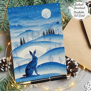 Peut inclure: Une carte imprimée d'art de paysage d'hiver bleu et blanc avec un lièvre, des collines enneigées et une pleine lune. La carte est étiquetée "Téléchargement instantané Carte imprimable 5x7". Un stylo noir et une enveloppe brune sont également visibles.