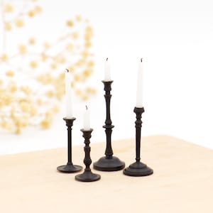 Könnte beinhalten: Vier schwarze Miniatur-Kerzenhalter mit weißen Kerzen. Die Kerzenhalter haben unterschiedliche Höhen und Designs, mit einer klassischen, verzierten Ästhetik. Die Kerzen sind unbeleuchtet, mit sichtbaren Dochten. Der Hintergrund ist eine helle Holzoberfläche und ein weißer Hintergrund.