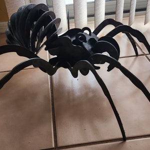 Könnte beinhalten: Eine große schwarze Spinnen-Skulptur aus Metall mit acht langen Beinen. Die Spinne ist mit ausgebreiteten Beinen posiert, als würde sie kriechen. Die Skulptur besteht aus vielen zusammengeschweißten Metallteilen.