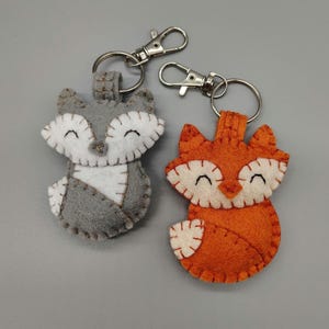 May include: Two felt fox keychains, one gray and one orange, with white accents and smiling faces. The keychains are attached to silver key rings.