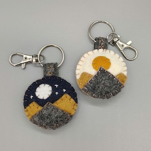 May include: Two circular felt keychains with metal lobster claw clasps. One features a dark blue night sky with a white moon, stars, and mustard yellow mountains. The other shows a white sky with a mustard yellow sun and grey mountains.