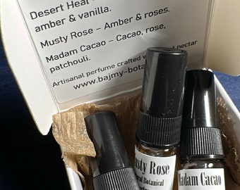 May include: Three small glass bottles of perfume in a white box. The bottles are labeled "Desert Heat", "Musty Rose", and "Madam Cacao". The box is decorated with brown paper and the text "BAJMY Did you come of your own free will, or at another's bidding?"