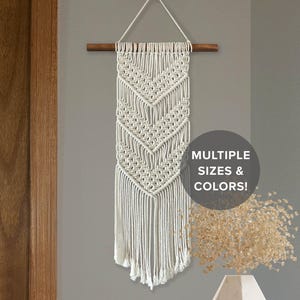 May include: Cream-colored macrame wall hanging with a geometric chevron design, suspended from a wooden dowel. The text "MULTIPLE SIZES & COLORS!" is displayed on the right side of the image. This decorative piece adds a bohemian touch to any space.