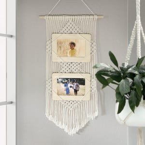 May include: A macrame wall hanging with two wooden picture frames. The frames hold photographs and are suspended from a wooden dowel. The macrame is made of off-white cotton cord, creating a bohemian aesthetic. The wall hanging is a decorative home decor item.