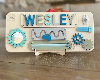 May include: A wooden activity board with a blue, green, and white color scheme. The board features a zipper, a spinning gear, a sliding bead, a wavy track with a car, and a mirror. The board is personalized with the name "WESLEY".