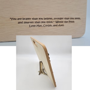 May include: A wooden cutting board with a stand, featuring an engraved quote from Winnie the Pooh: "You are braver than you believe, stronger than you seem, and smarter than you think." The board is signed "Love Mat, Corbin, and Axel."