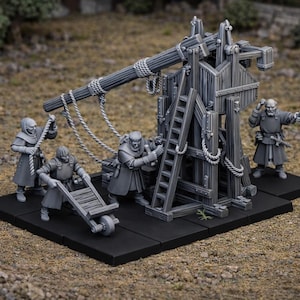 Puede incluir: Maqueta gris de un trabuquete con figuras, cuerdas y escaleras. El modelo está sobre una base negra, con figuras vestidas con atuendo medieval. El fondo es una escena exterior borrosa.
