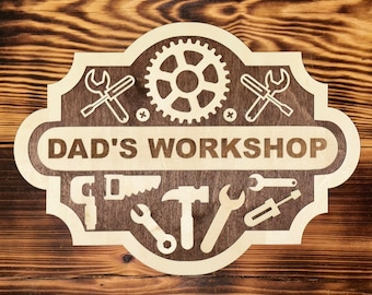 May include: Wooden sign with the text "DAD'S WORKSHOP" and tool illustrations. The sign features a light-colored border and tools like wrenches, a saw, and a hammer. The background is a dark wood grain.