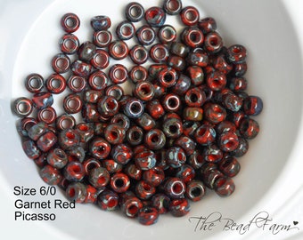 May include: A pile of red, blue, and gray 6/0 size glass seed beads with a mottled, marbled, or Picasso finish. The beads are in a white bowl. The text "Size 6/0 Garnet Red Picasso" is visible below the bowl.