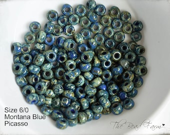 May include: A close-up of a pile of blue and green glass beads. The beads are size 6/0 Montana Blue Picasso.