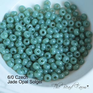 May include: A collection of 6/0 Czech Jade Opal Solgel beads. The beads are a light teal color and have a rounded shape with a hole through the center. The text "The Bead Farm" is in the bottom right corner.