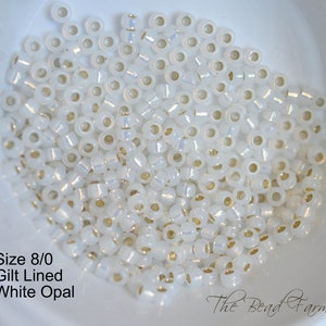 May include: A close-up of a bowl filled with white opal beads with gold-lined edges. The beads are size 8/0. The text "Size 8/0 Gilt Lined White Opal" is visible below the bowl. The text "The Bead Farm" is visible in the bottom right corner of the image.