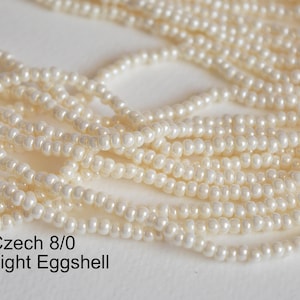 May include: A close-up of a strand of white Czech 8/0 seed beads in the color Light Eggshell.