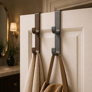 May include: Two metal over-the-door hooks in brown and gray. A beige towel hangs from the brown hook, and a brown purse hangs from the gray hook. The hooks are mounted on a white door.