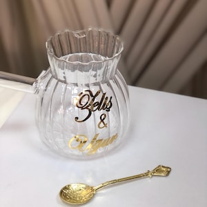 Op de afbeelding: Een heldere glazen Turkse koffiepot met een gouden inscriptie die "Selis & Ugur" luidt. Een gouden lepel met een sierlijk handvat is ook afgebeeld.