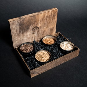 May include: A wooden box with a lid, containing four small black containers filled with different types of wood shavings. The box has a label with the text "Selvage" on it.
