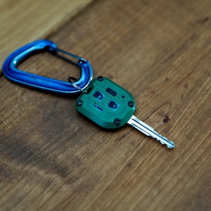 May include: A metallic green car key fob with a square shape and four black screws at the corners, featuring a central button interface with lock and unlock icons. The fob is attached to a bright blue metal carabiner by a small silver ring.