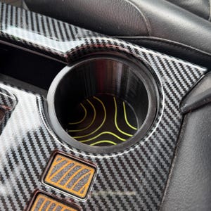 May include: A black circular car cup holder insert featuring a yellow topographic map line pattern. The insert is positioned within a vehicle center console that has a glossy black carbon fiber style finish and orange rectangular accent buttons.