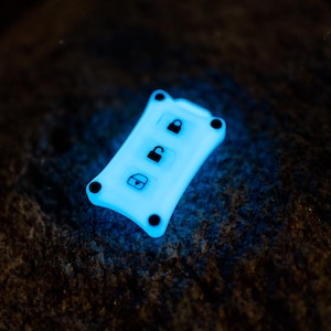 Puede incluir: Una funda rectangular de silicona que brilla en la oscuridad y emite una luz azul brillante. La funda presenta tres iconos negros que representan un candado cerrado, un candado abierto y un maletero, con cuatro pequeños acentos circulares negros.